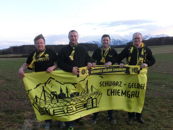 Vier Männer in schwarzen Fanclub-Shirts halten auf einer Wiese ein gelbes Banner des Schwarz-Gelben Chiemgau, dahinter Berge.