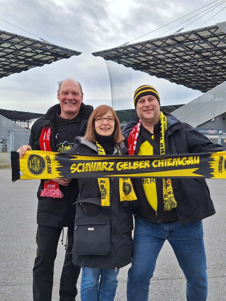 Zwei Männer und eine Frau halten vor dem Stadion einen Schal des Schwarz-Gelben Chiemgau.
