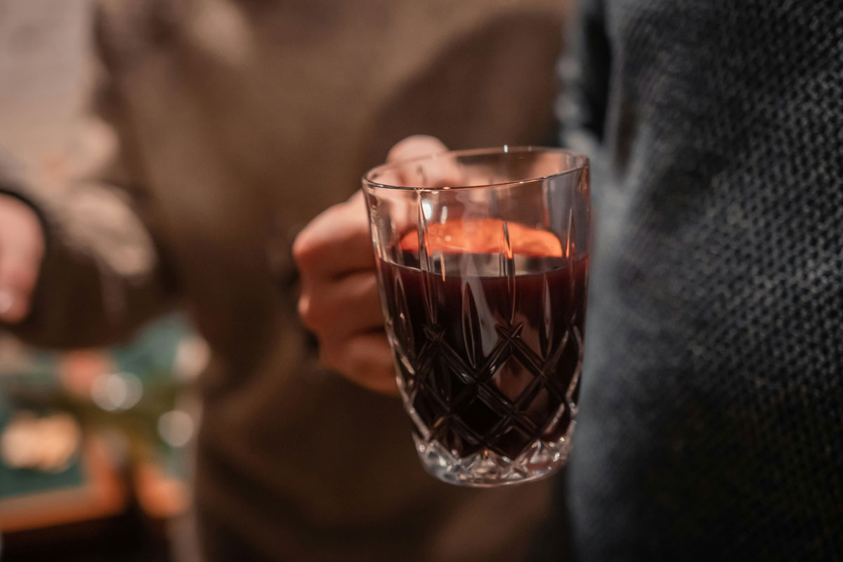 Hand hält ein geschliffenes Glas mit dampfendem rotem Glühwein vor unscharfem Hintergrund.