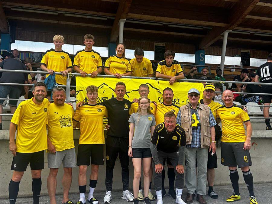 Rund 15 Spieler und Fans in gelben Trikots stehen vor einer überdachten Tribüne mit Fanclub-Banner.