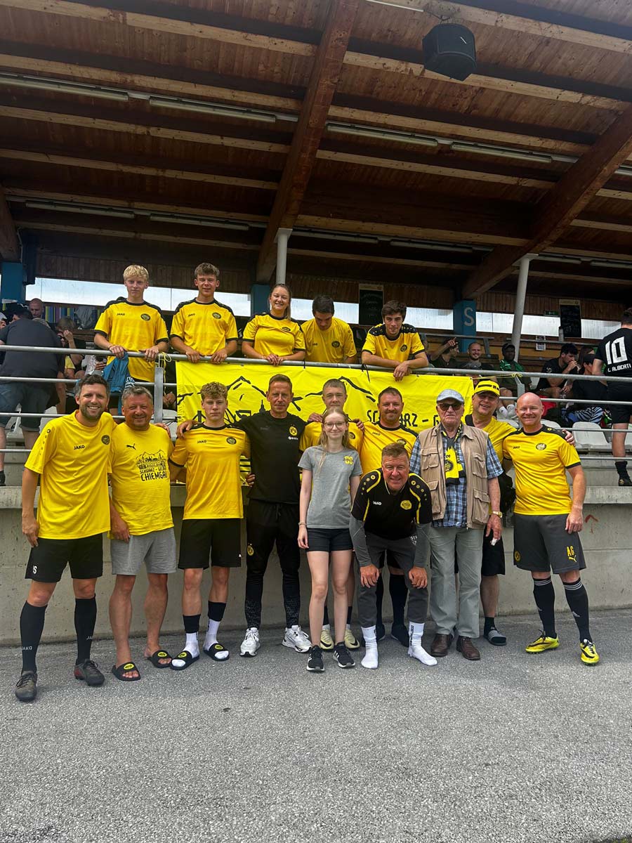 Rund 15 Spieler und Fans in gelben Trikots stehen vor einer überdachten Tribüne mit Fanclub-Banner.