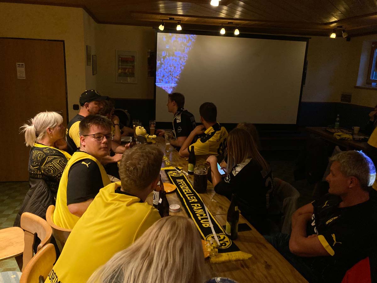 Mitglieder in BVB-Trikots sitzen an Holztischen in einem Raum vor einer Leinwand.
