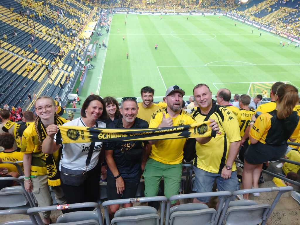 Gruppe von Mitgliedern in gelben BVB-Trikots hält auf der Tribüne einen Schal hoch, dahinter das Spielfeld.