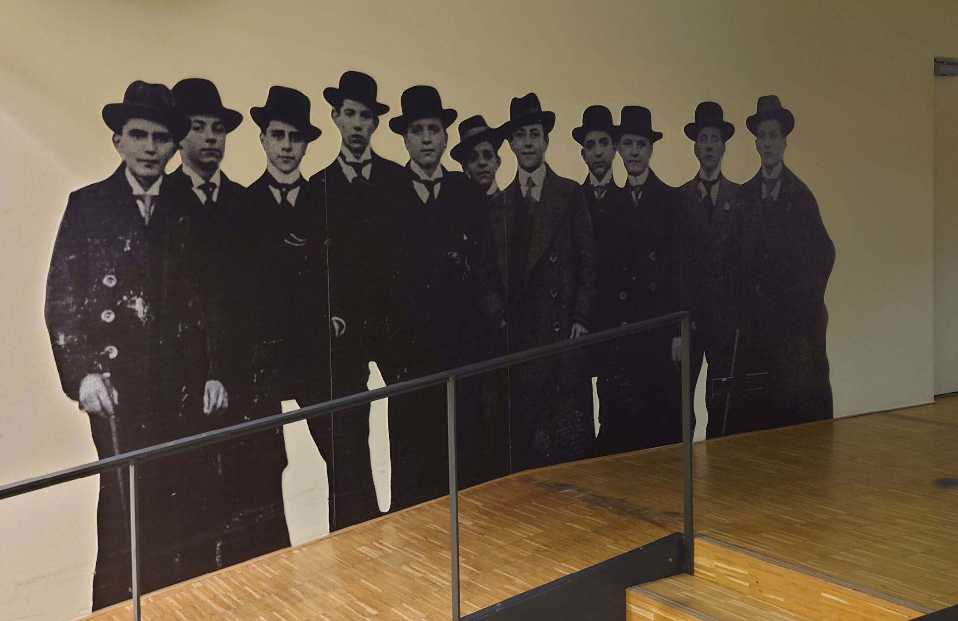 Wandbild mit lebensgroßen Silhouetten von Männern in Anzügen und Hüten hinter einem Geländer.