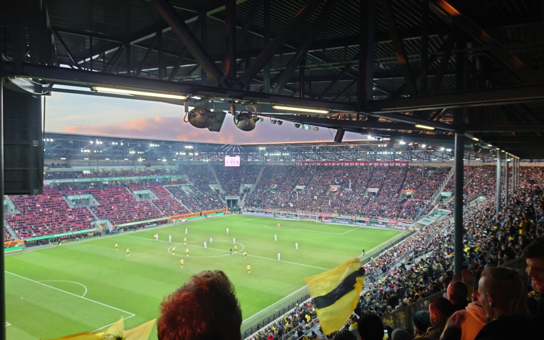 Blick aus dem Gästeblock unter dem Stadiondach auf das beleuchtete Spielfeld und rote Sitzreihen.