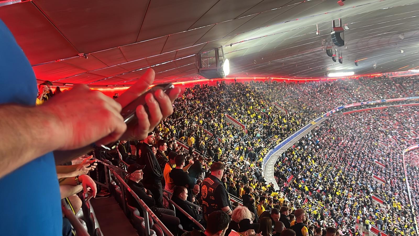 Blick über eine Hand hinweg auf den vollen gelben Gästeblock unter rot beleuchtetem Arenadach.