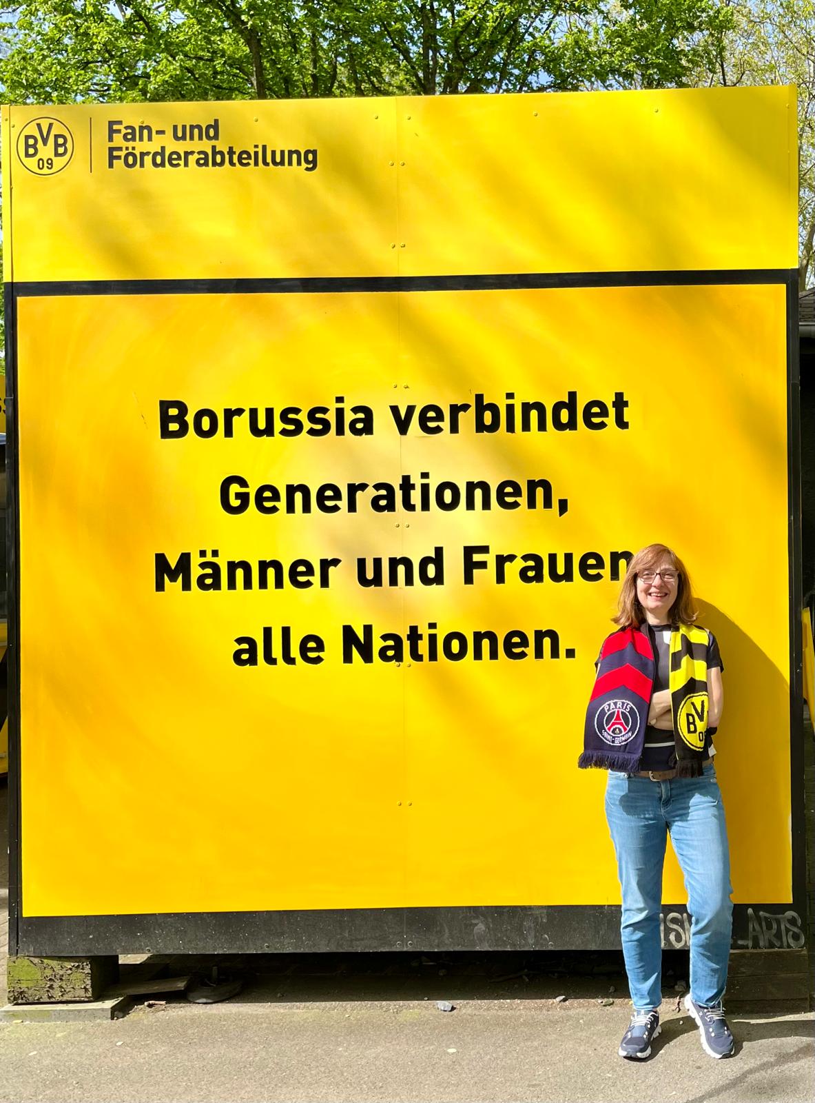 Frau mit BVB-Schal steht vor einer gelben Tafel mit der Aufschrift Borussia verbindet Generationen, Männer und Frauen, alle Nationen.