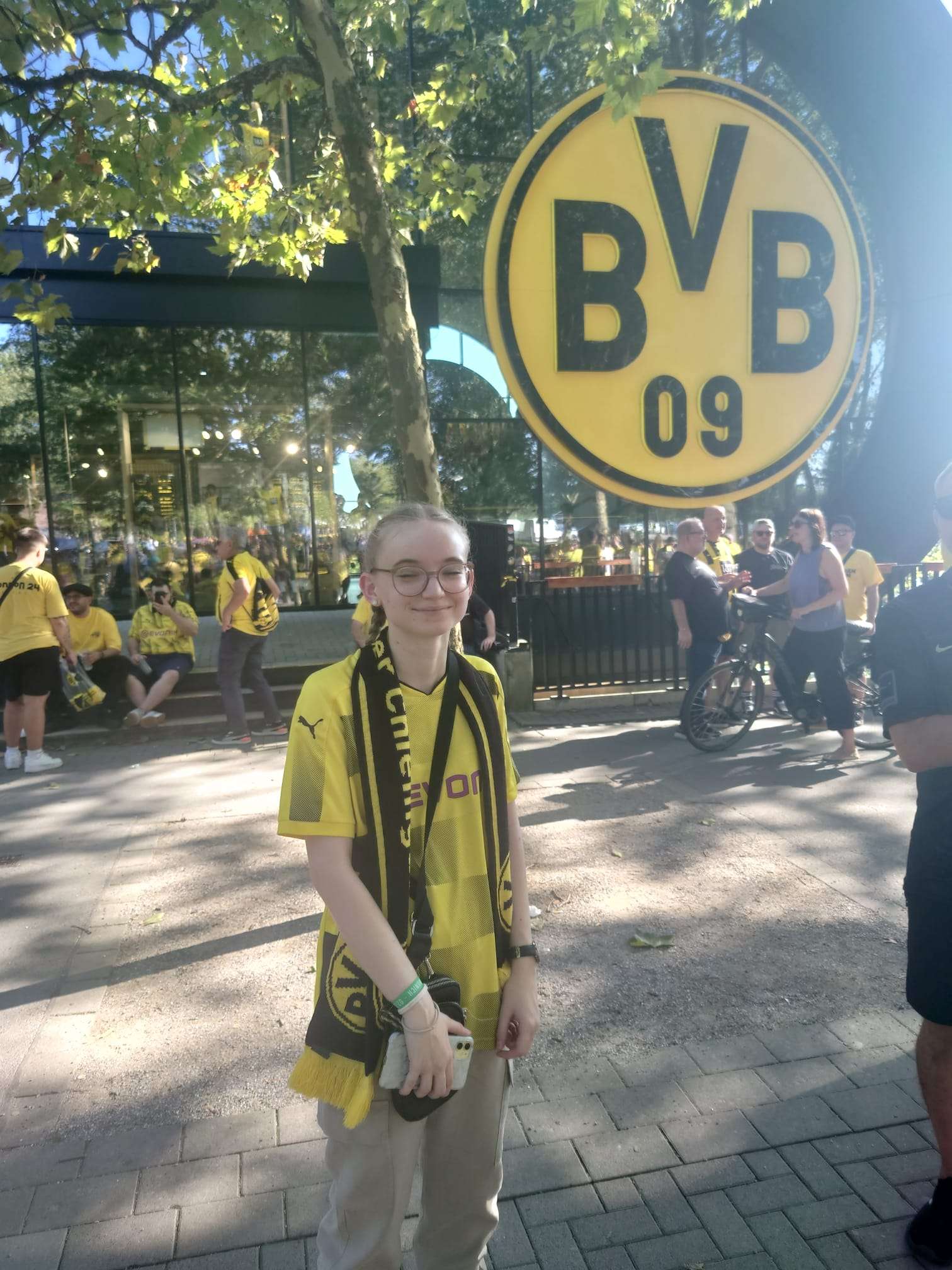 BVB - Eintracht Frankfurt Junger Mann im gelben BVB-Trikot mit Schal steht unter Bäumen vor einem großen BVB-Logo.