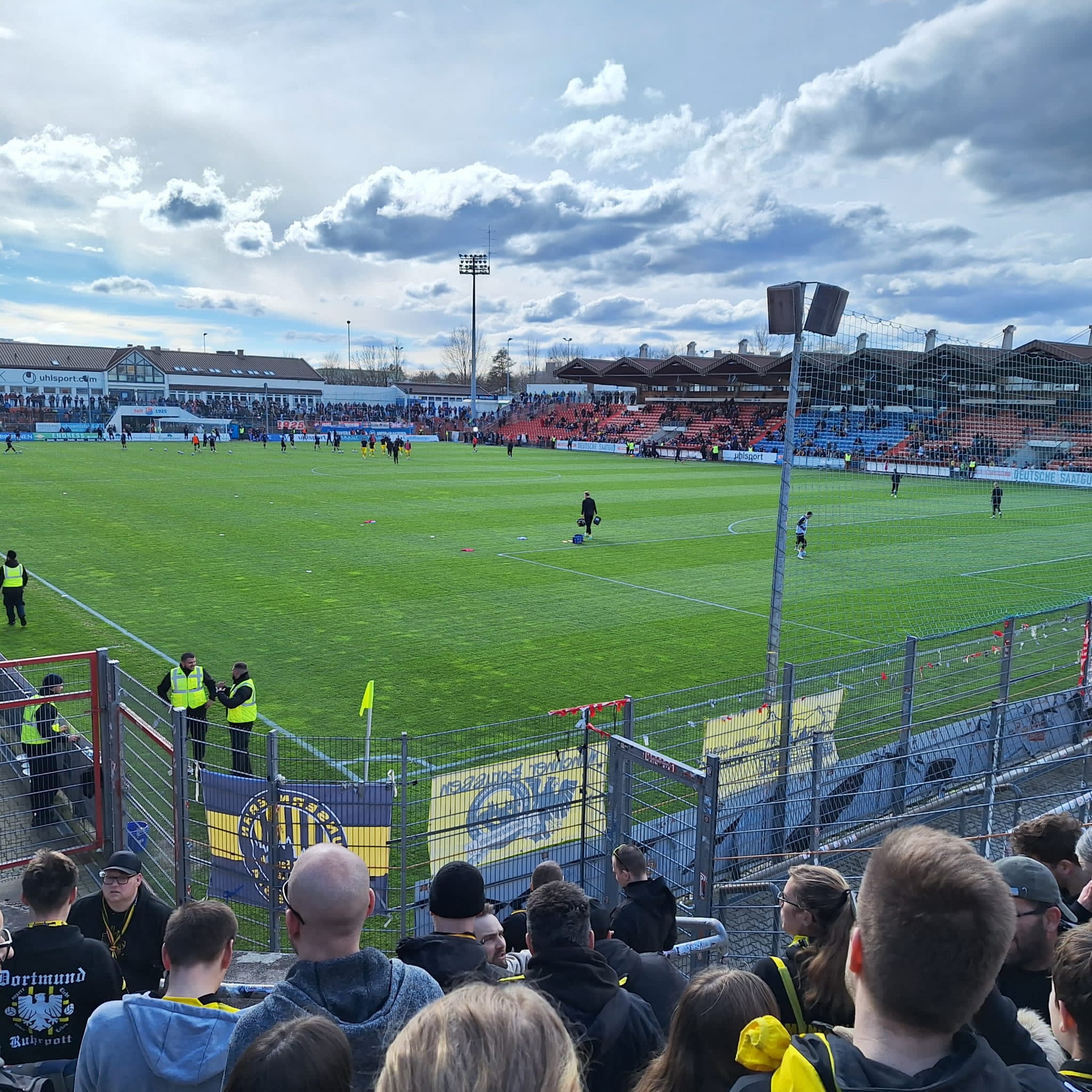 Blick über Fans und Zaun auf das sonnige Spielfeld, gegenüber eine Tribüne mit blauen Sitzen und Flutlichtmasten.