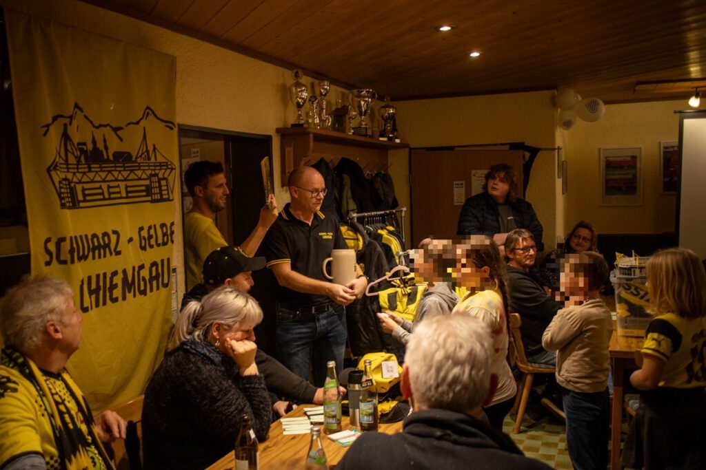 Mitglieder sitzen dicht an Tischen in einem holzvertäfelten Raum, links hängt das gelbe Fanclub-Banner.