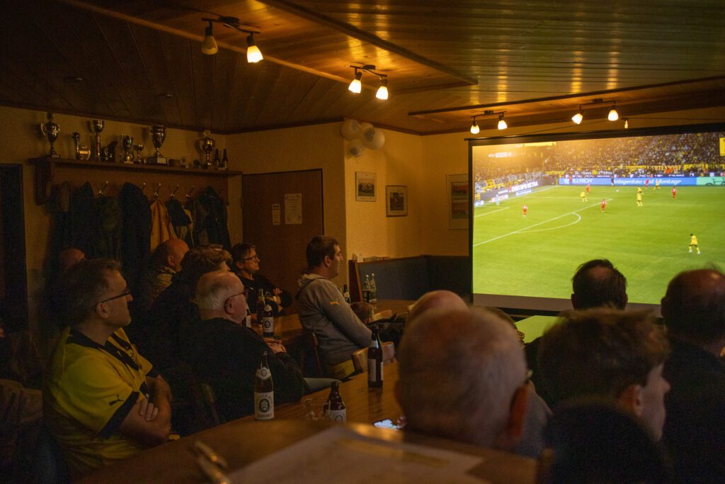 Mitglieder sitzen im abgedunkelten Sportheim und blicken auf eine Leinwand mit laufendem Fußballspiel.