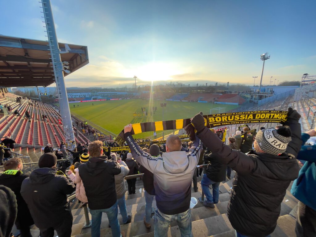 Fans recken auf der Tribüne einen schwarz-gelben Schal in die Höhe, im Gegenlicht die tiefstehende Sonne.