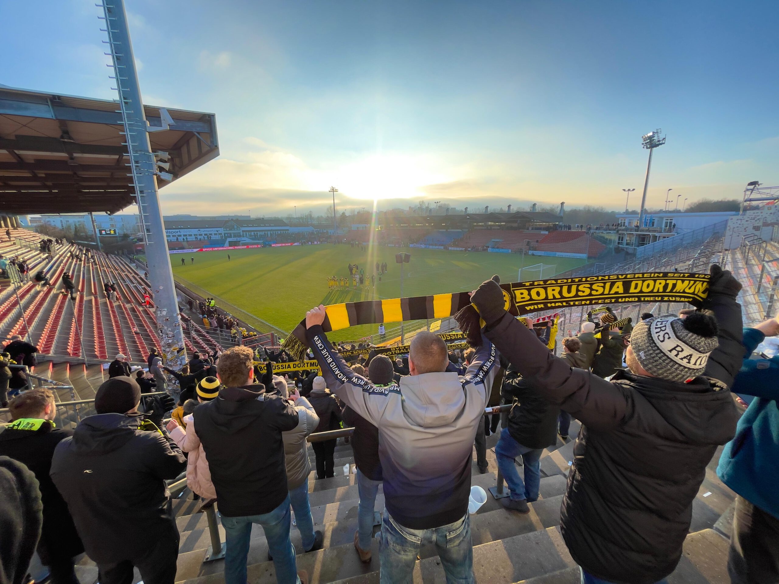 Fans recken auf der Tribüne einen schwarz-gelben Schal in die Höhe, im Gegenlicht die tiefstehende Sonne.