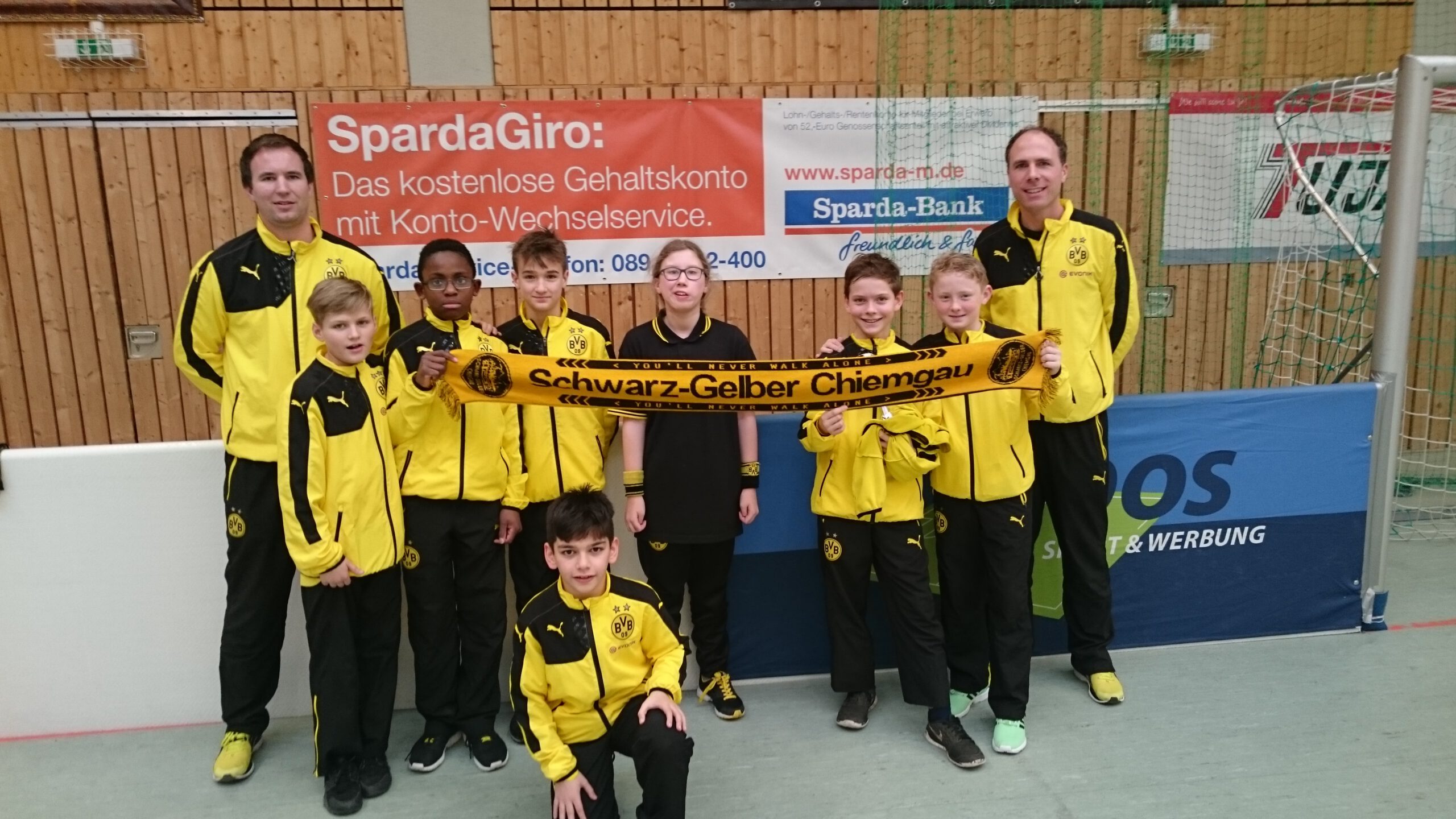 Sieben Kinder und zwei Erwachsene in schwarz-gelben Trainingsanzügen halten in einer Sporthalle einen Fanclub-Schal.