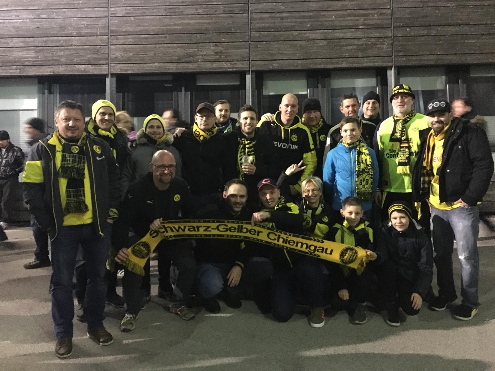 Rund 15 Mitglieder in Winterjacken mit BVB-Schals stehen und hocken vor einer Holzfassade, vorne ein Fanclub-Schal.