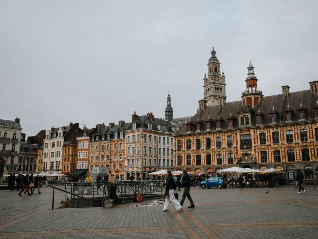Weiter Platz mit Passanten vor einer Reihe historischer Backsteinfassaden und einem Turm unter grauem Himmel.