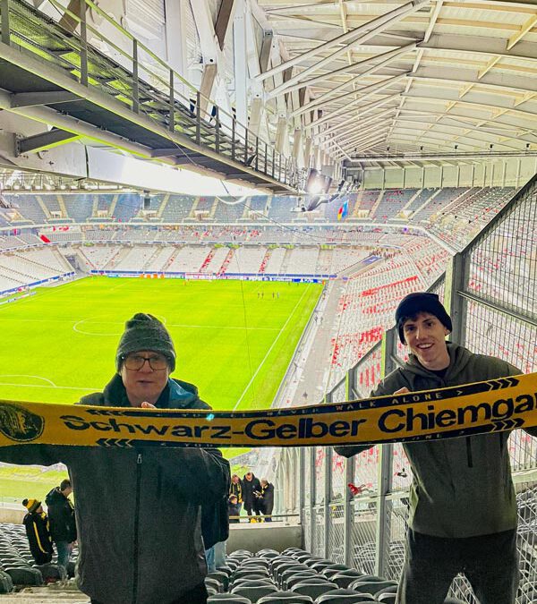 Zwei junge Fans halten einen Schal des Schwarz-Gelben Chiemgau, hinter ihnen das Spielfeld und leere Sitzreihen.