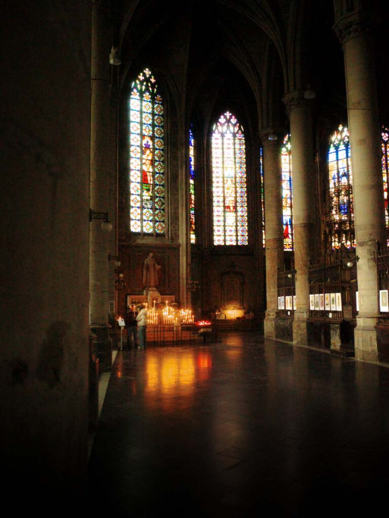 Dämmriges Kirchenschiff mit Säulen, bunten Glasfenstern und Kerzenlicht, das sich im Boden spiegelt.