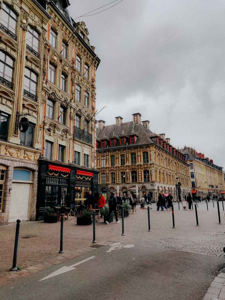 Gepflasterte Straße mit Pollern zwischen verzierten historischen Fassaden unter bewölktem Himmel.