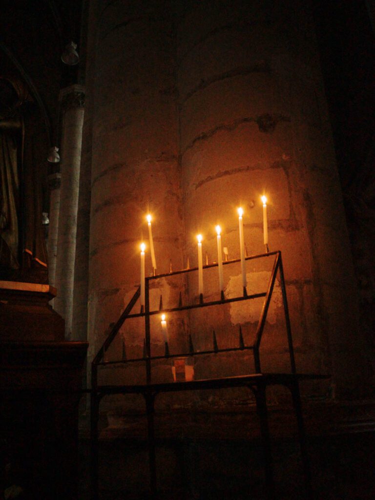 Kerzenständer mit mehreren brennenden Kerzen vor einem steinernen Pfeiler im warmen Licht.
