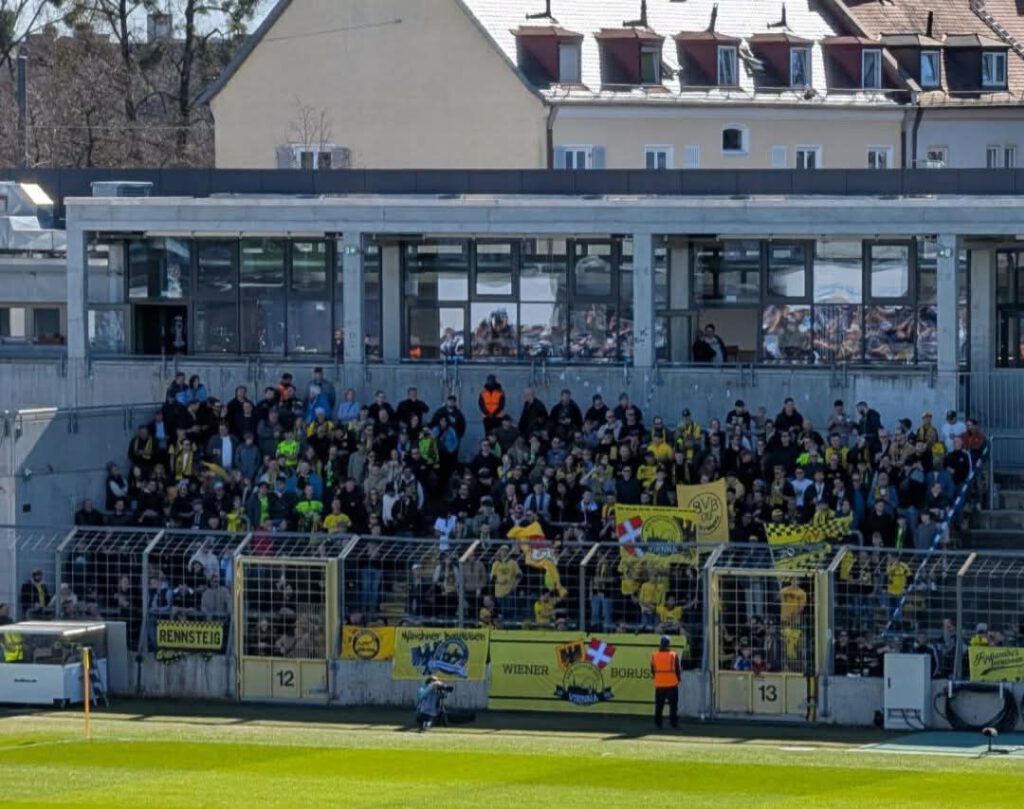 Volle überdachte Tribüne mit Fans in Schwarz-Gelb, am Zaun davor hängen Fahnen und Banner, vorne der Rasen.