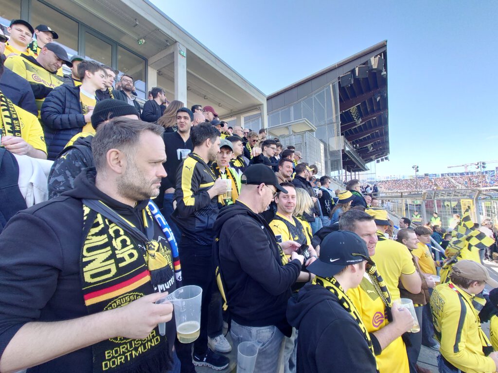 Mann mit Bierbecher und BVB-Schal im dicht gefüllten Gästeblock unter blauem Himmel.