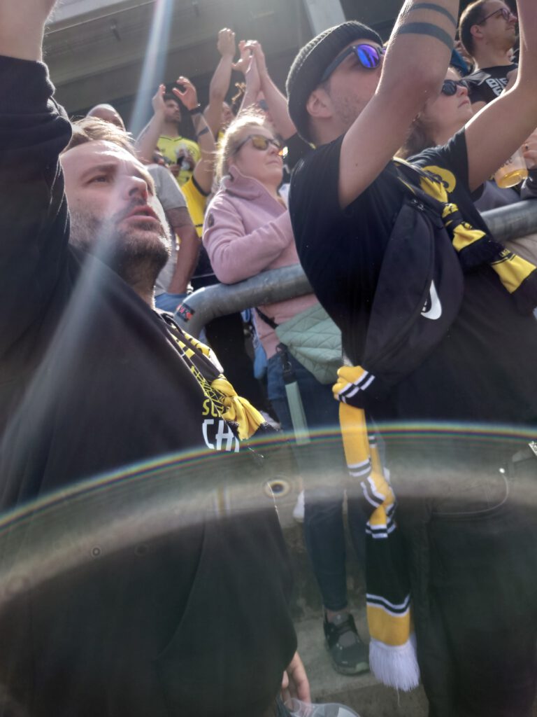 Fans in BVB-Kleidung jubeln im Block mit erhobenen Armen, vorne ein Bierbecher.