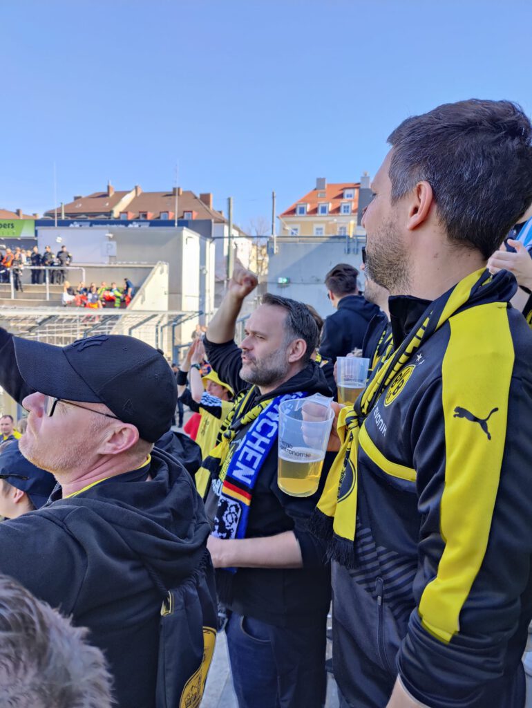 Zwei Fans in BVB-Jacken, einer mit Bierbecher, schauen im Block Richtung Spielfeld.