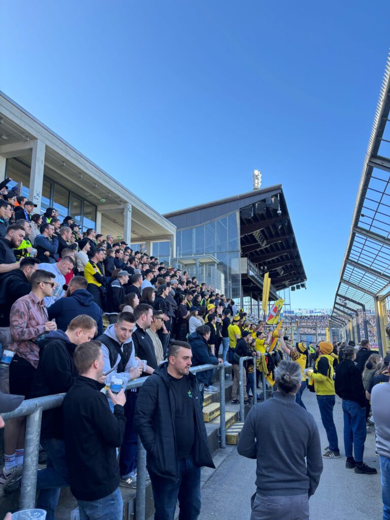 Blick die Stufen hinauf auf einen vollen Block mit Fans in Schwarz-Gelb, darüber blauer Himmel.