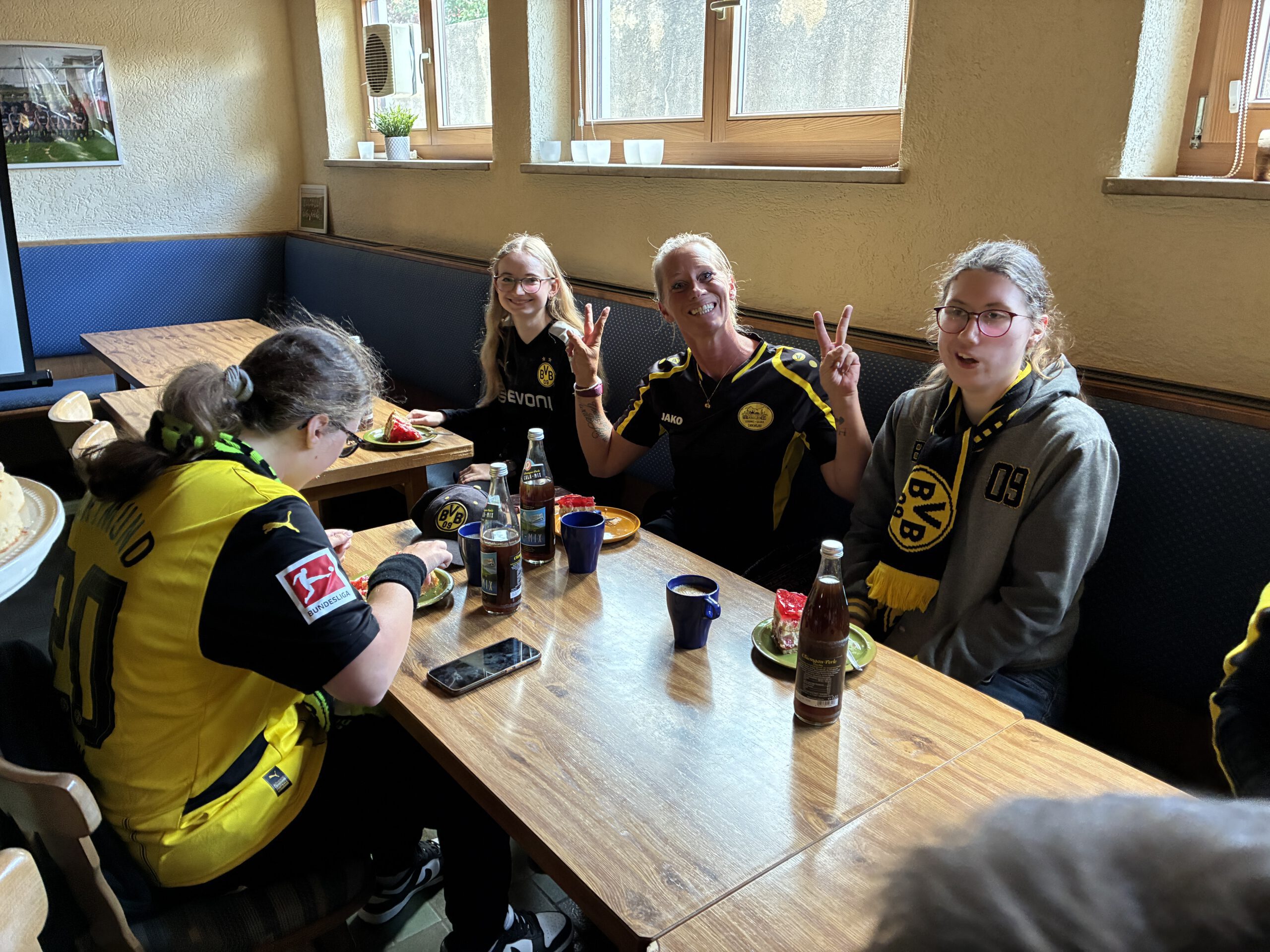 Sommerfest 2025 Drei Mitglieder in BVB-Kleidung sitzen am Tisch und zeigen das Victory-Zeichen, vorne eine weitere Person.