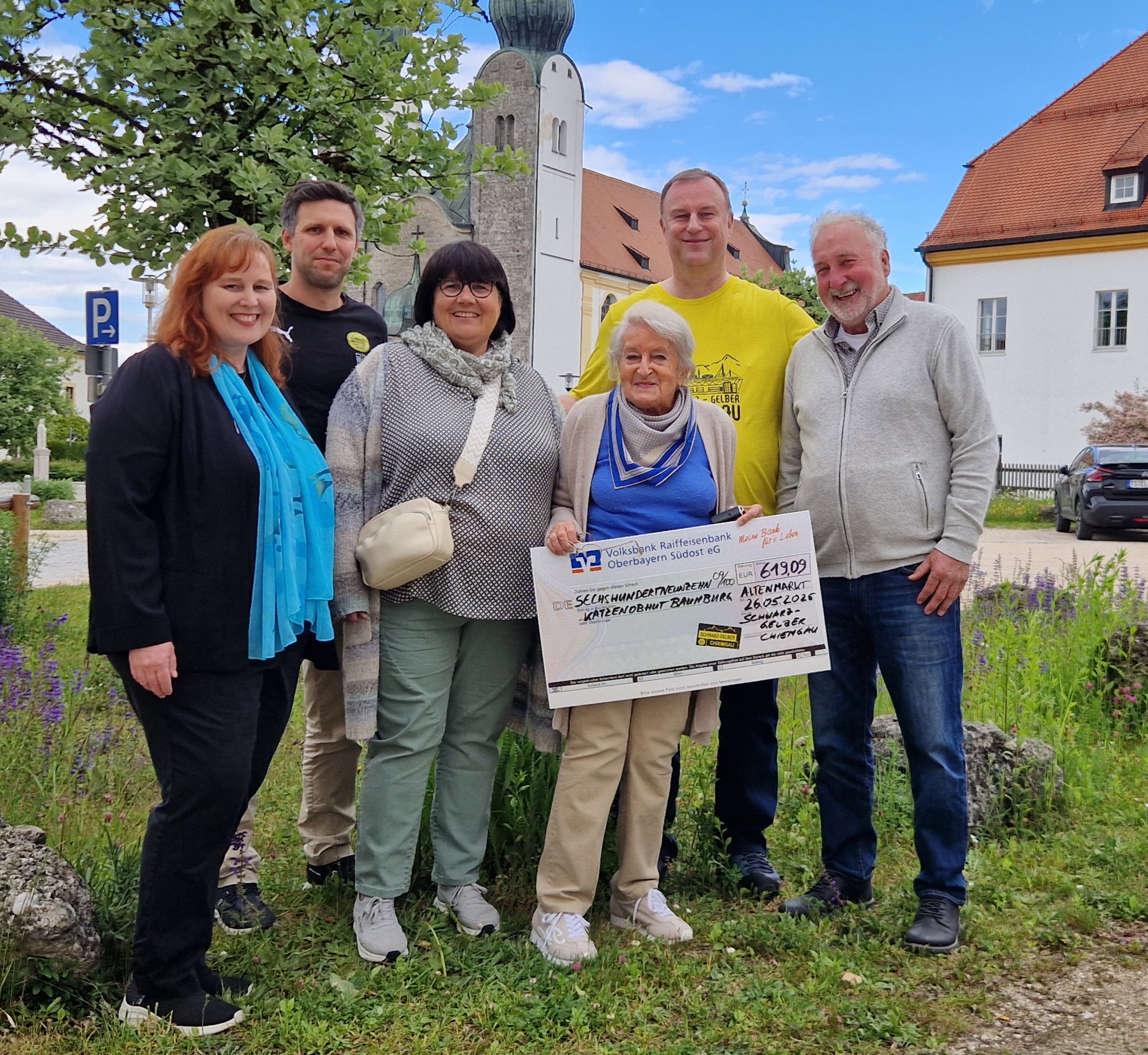 Sechs Personen stehen vor einer Kirche auf einer Wiese, eine Frau hält einen Spendenscheck.