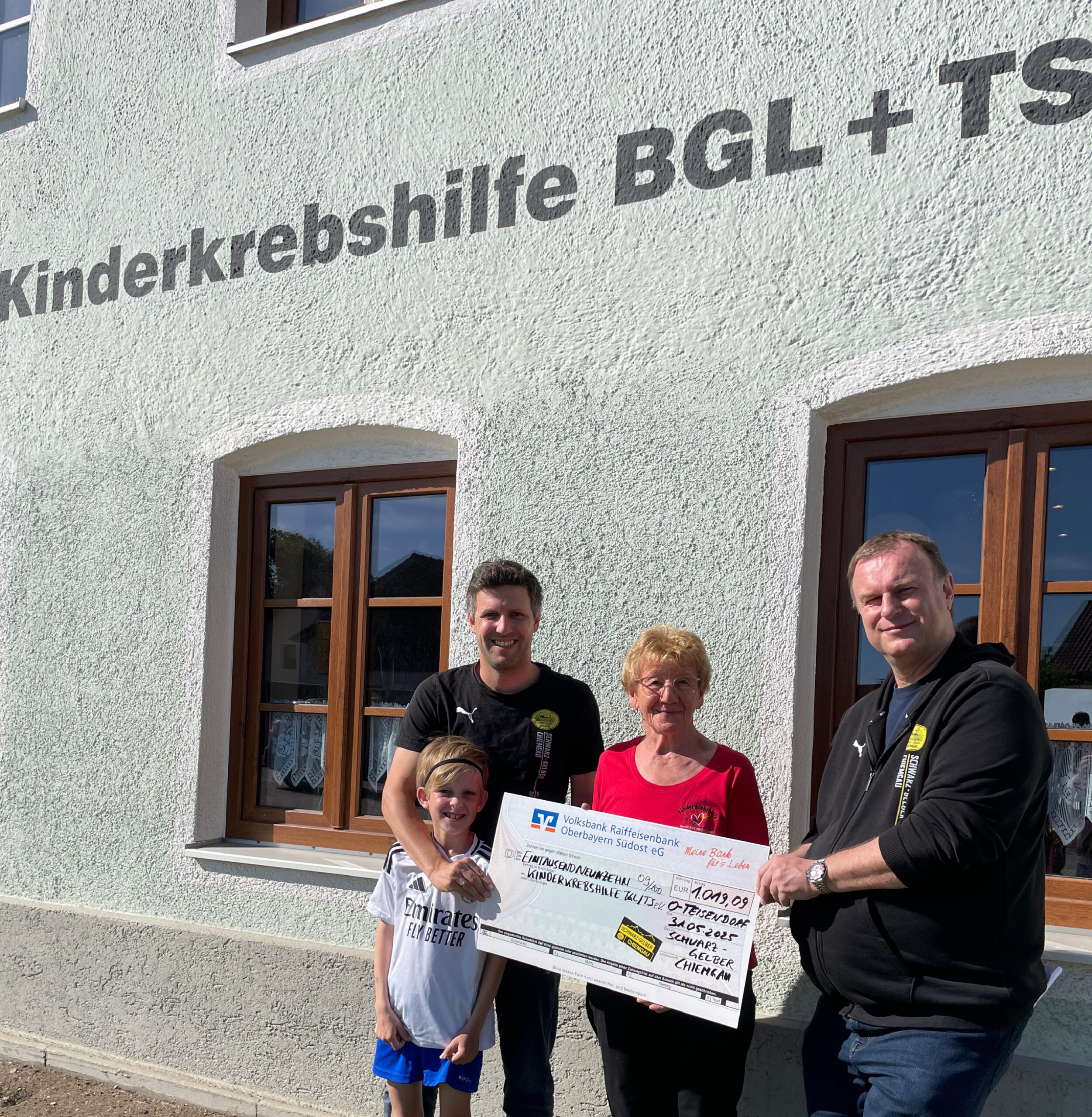 Zwei Männer, eine Frau und ein Kind halten einen Spendenscheck vor einem weißen Haus mit der Aufschrift Kinderkrebshilfe BGL + TS.