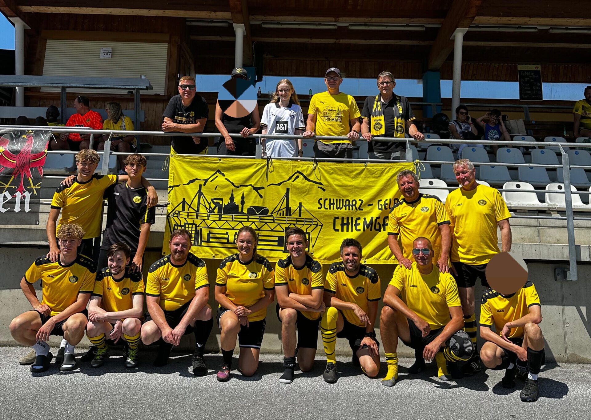 Rund 15 Spieler und Fans in gelben Trikots vor einem gelben Fanclub-Banner an einer überdachten Tribüne.