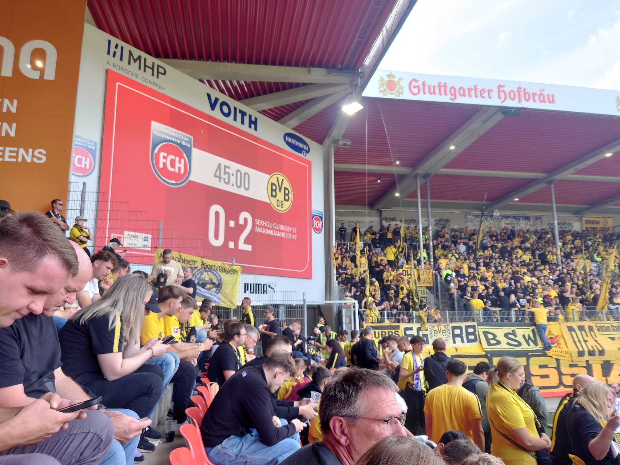 Rote Anzeigetafel zeigt zur Halbzeit 0:2, darunter Zuschauer auf der Tribüne und Fans im Gästeblock.