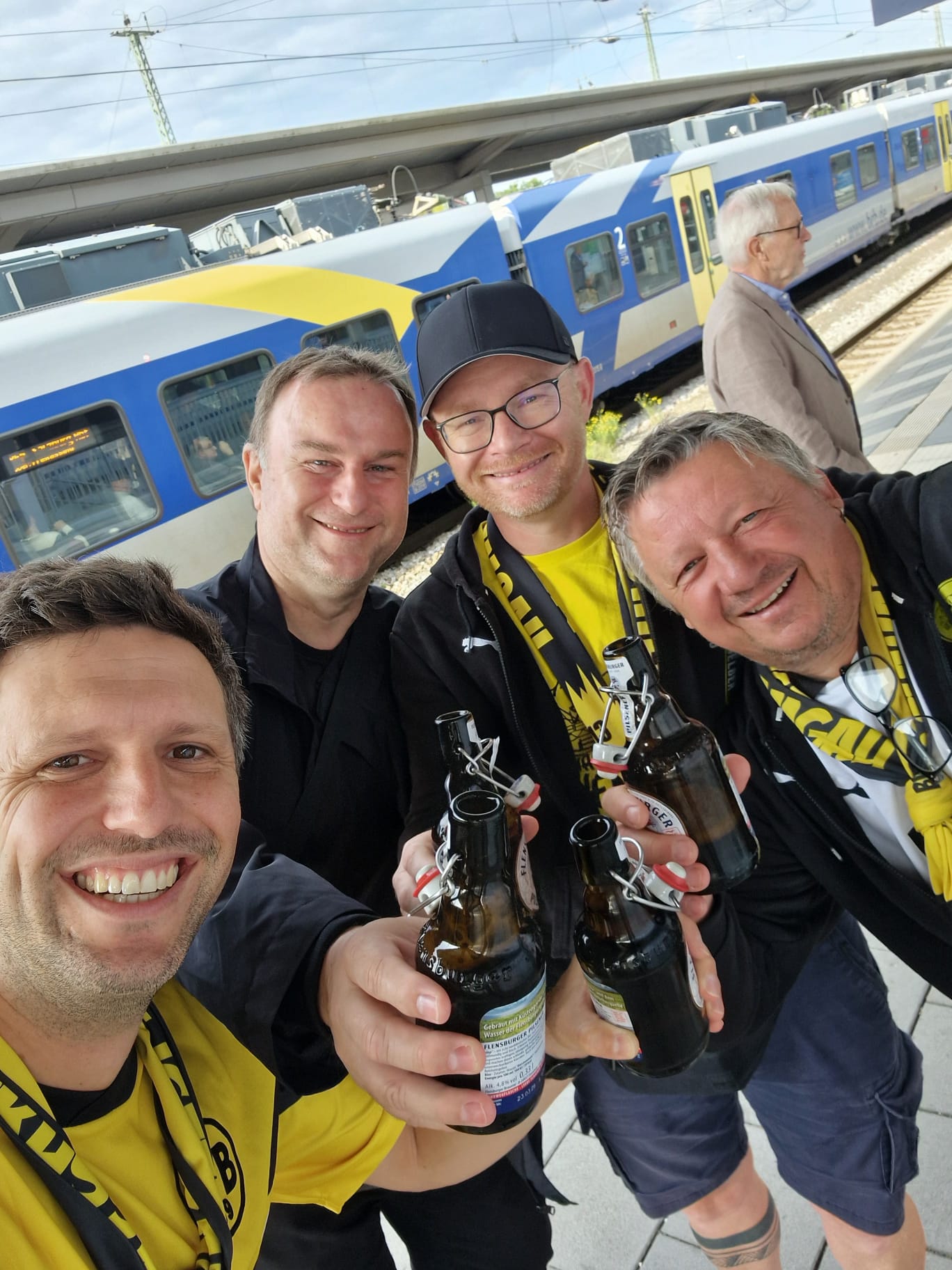 Vier gut gelaunte Männer in BVB-Kleidung mit Bierflaschen machen am Bahnsteig vor einem Zug ein Selfie.