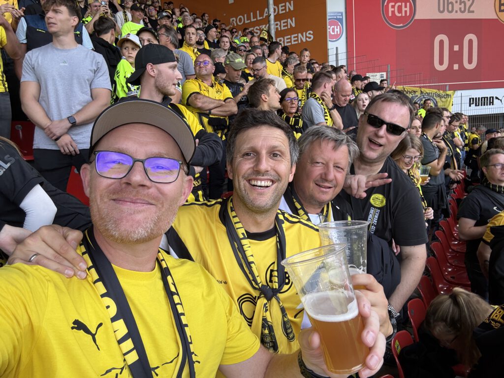 Fünf Mitglieder in gelben BVB-Shirts, einer mit Bierbecher, im vollen Gästeblock, im Hintergrund die Anzeige 0:0.