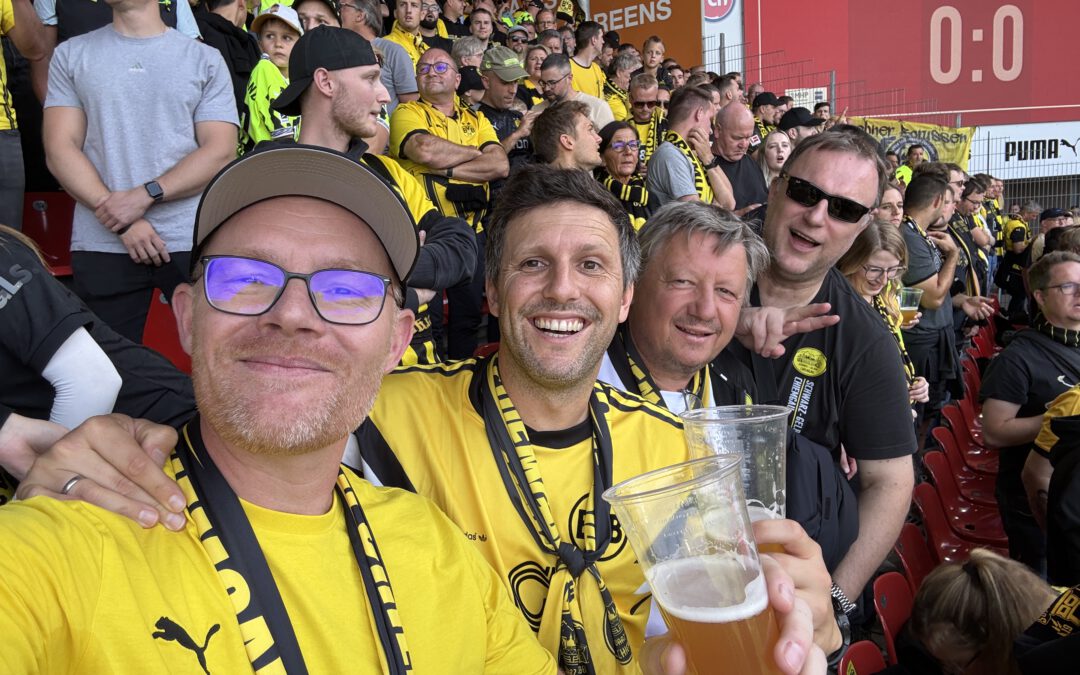 Fünf Mitglieder in gelben BVB-Shirts, einer mit Bierbecher, im vollen Gästeblock, im Hintergrund die Anzeige 0:0.
