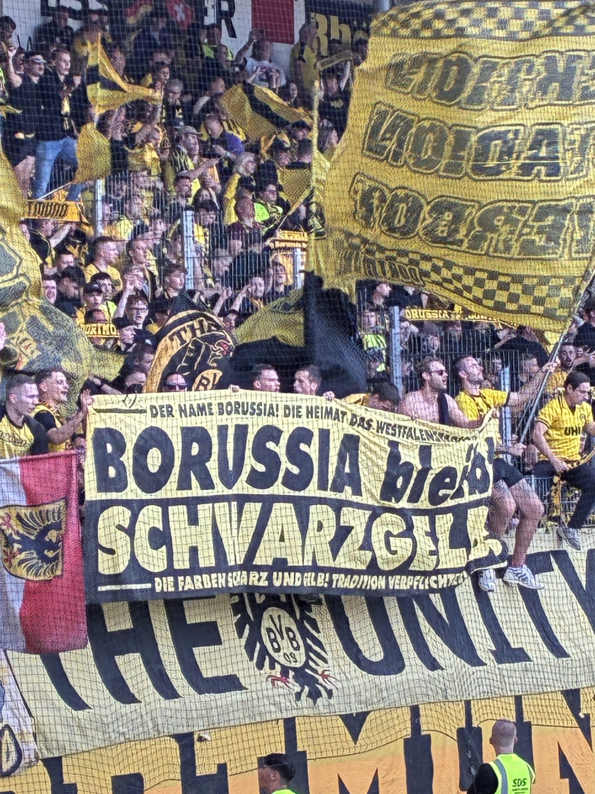 Hochformat: voller Gästeblock mit Fahnen und Spruchbändern in Schwarz-Gelb, darunter ein großes Borussia-Banner.