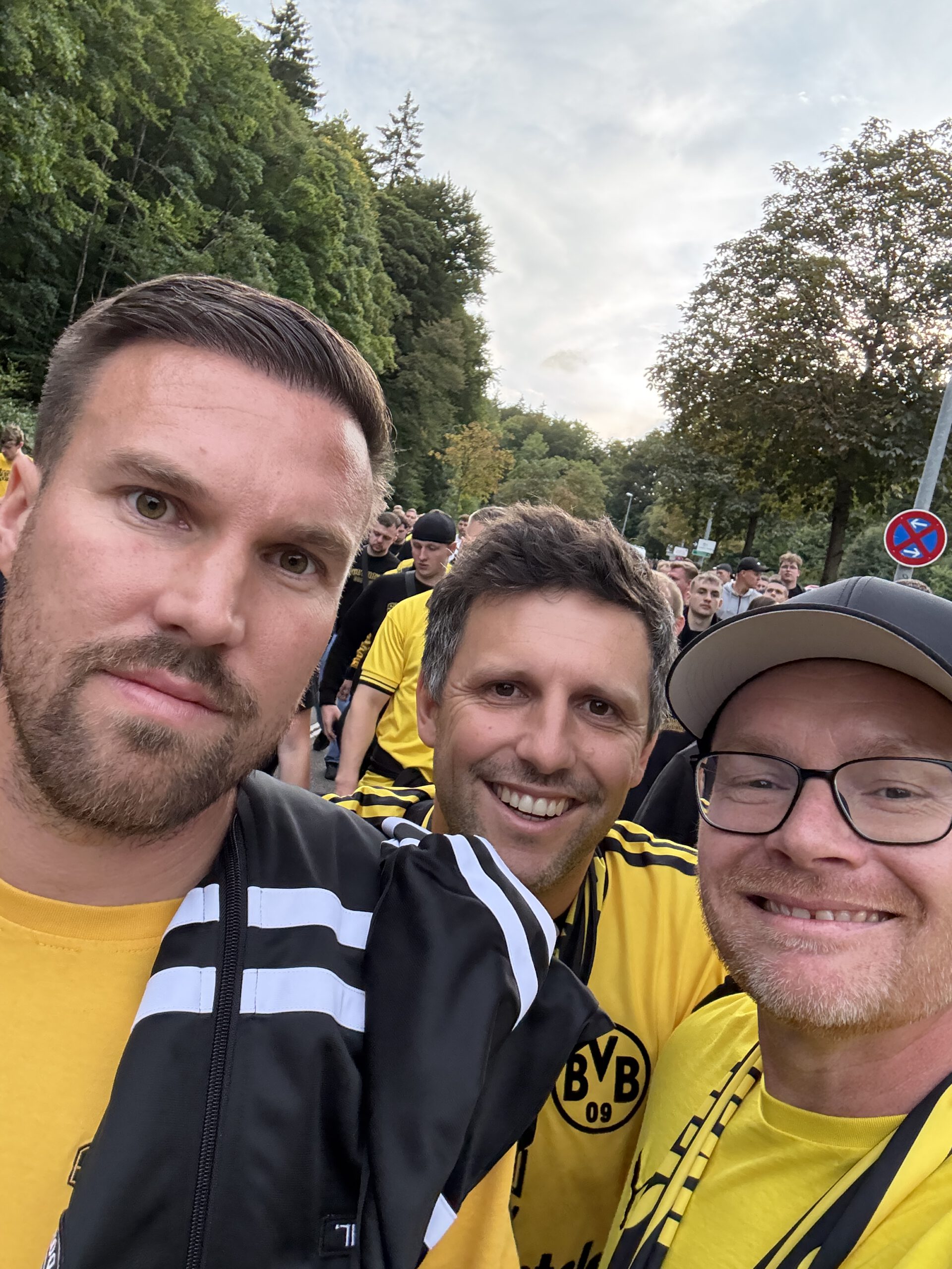 Drei Männer, zwei in gelben BVB-Shirts, machen ein Selfie unter Bäumen, dahinter weitere Fans.