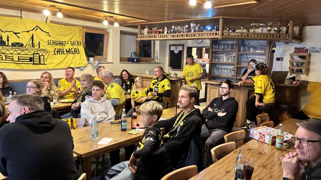 Mitglieder mit Schals sitzen an Tischen und schauen konzentriert nach vorn, an der Wand ein Fanclub-Banner.