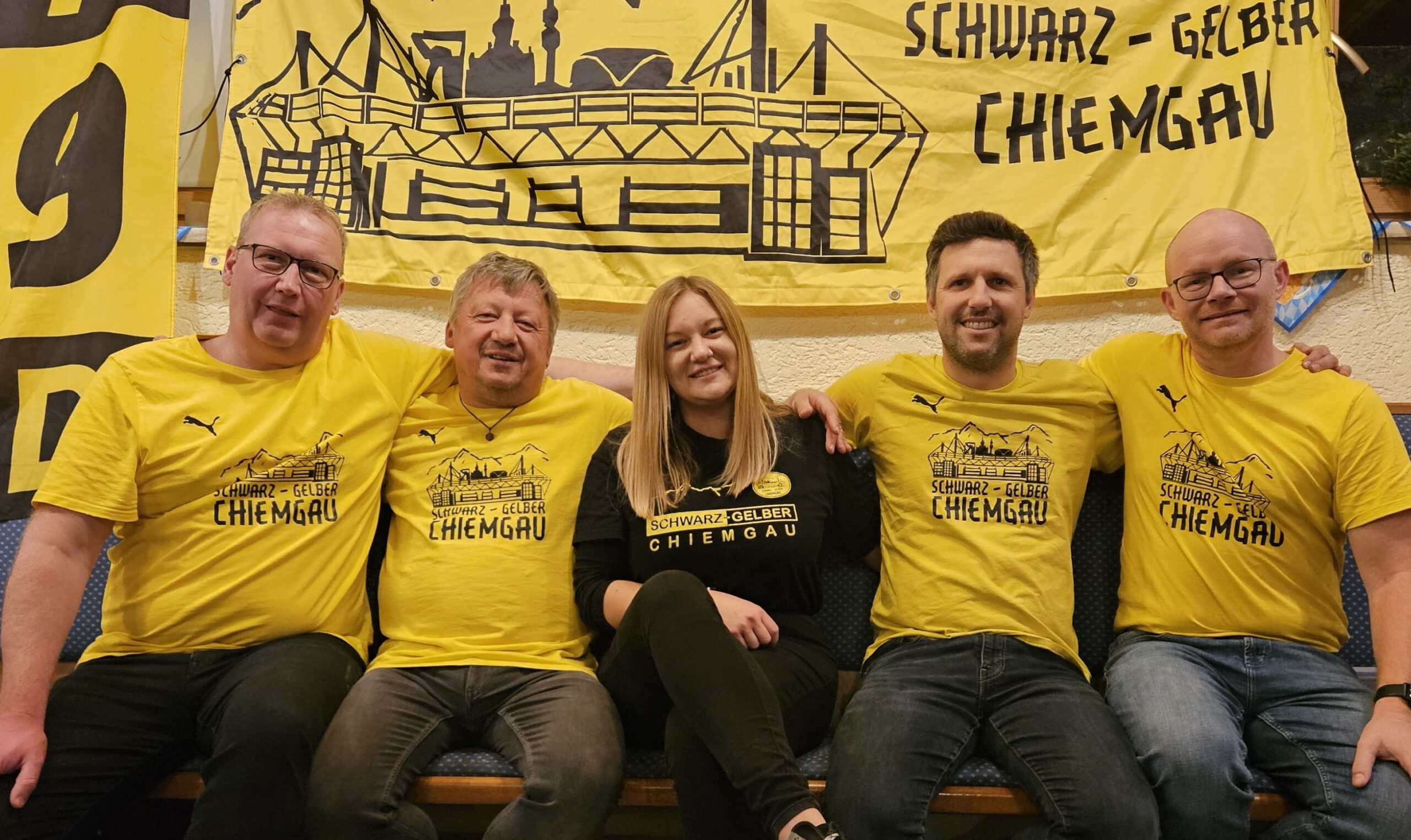 Fünf Personen in gelben und schwarzen Fanclub-Shirts sitzen nebeneinander vor dem gelben Fanclub-Banner.
