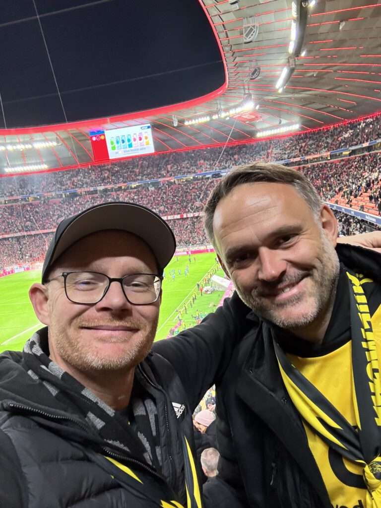 Zwei Männer mit Kappe und BVB-Schal machen ein Selfie, dahinter die roten Ränge der Allianz Arena.
