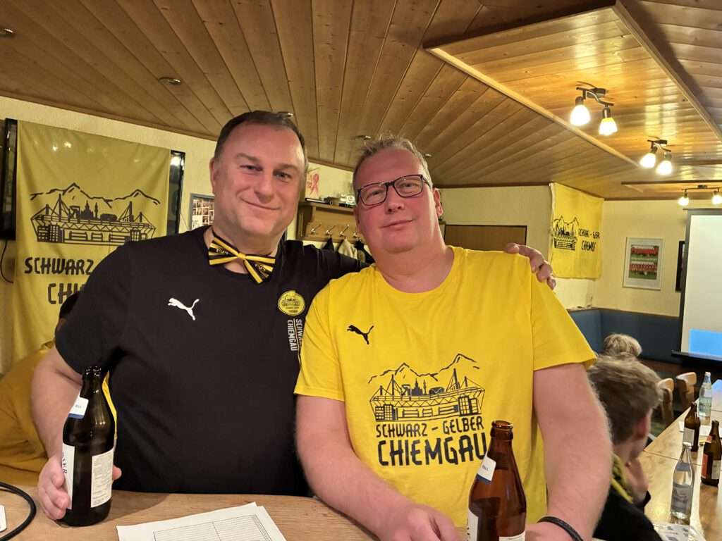 Zwei Männer in Fanclub-Shirts sitzen mit Bierflaschen an einem Tisch vor einem Fanclub-Banner.