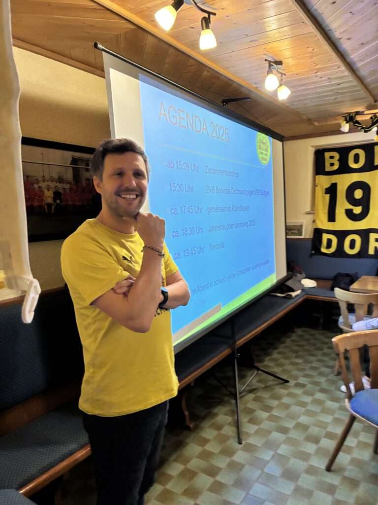 Mann im gelben Shirt steht neben einer Leinwand mit Präsentation in einem Vereinsraum.
