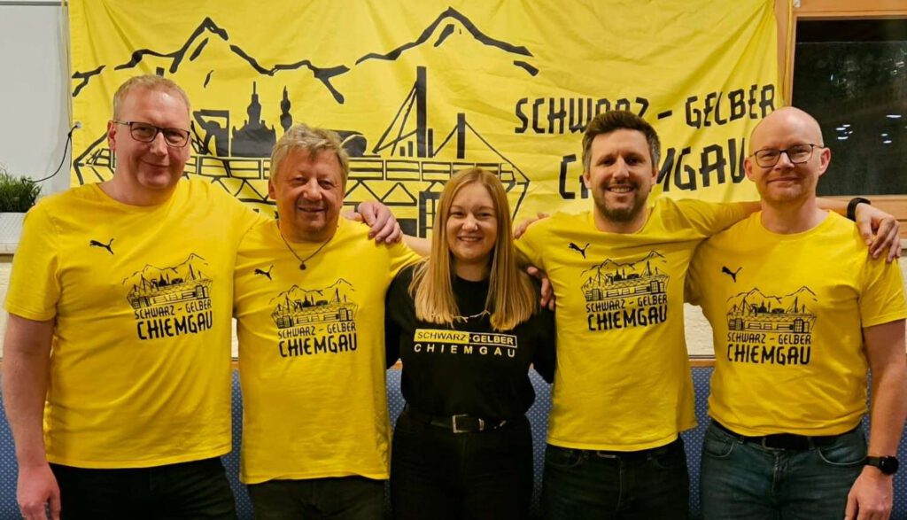 Fünf Personen in gelben Fanclub-Shirts stehen vor dem großen Banner des Schwarz-Gelben Chiemgau.
