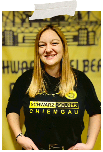Angeheftetes Foto: lächelnde Frau im schwarzen Fanclub-Shirt vor dem gelben Fanclub-Banner.
