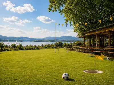 Sonnige Wiese mit Fußball und Torloch, Biergarten unter einem Baum, dahinter See und Berge.